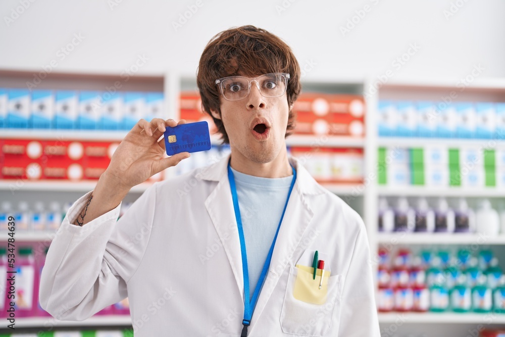 Hispanic young man working at pharmacy drugstore holding credit card scared and amazed with open mouth for surprise, disbelief face