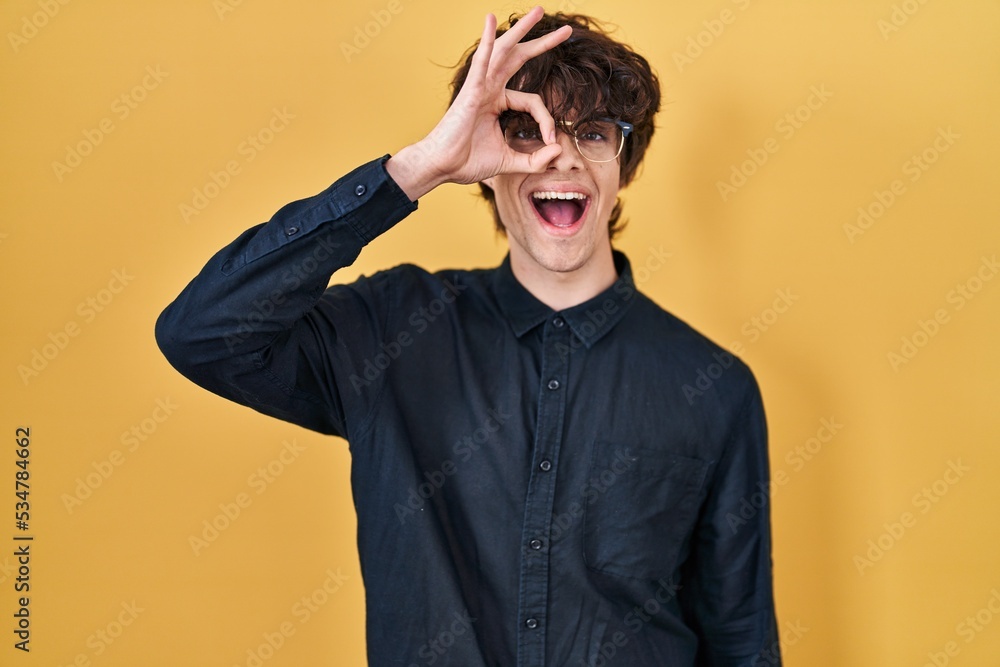 Young man wearing glasses over yellow background doing ok gesture with hand smiling, eye looking through fingers with happy face.