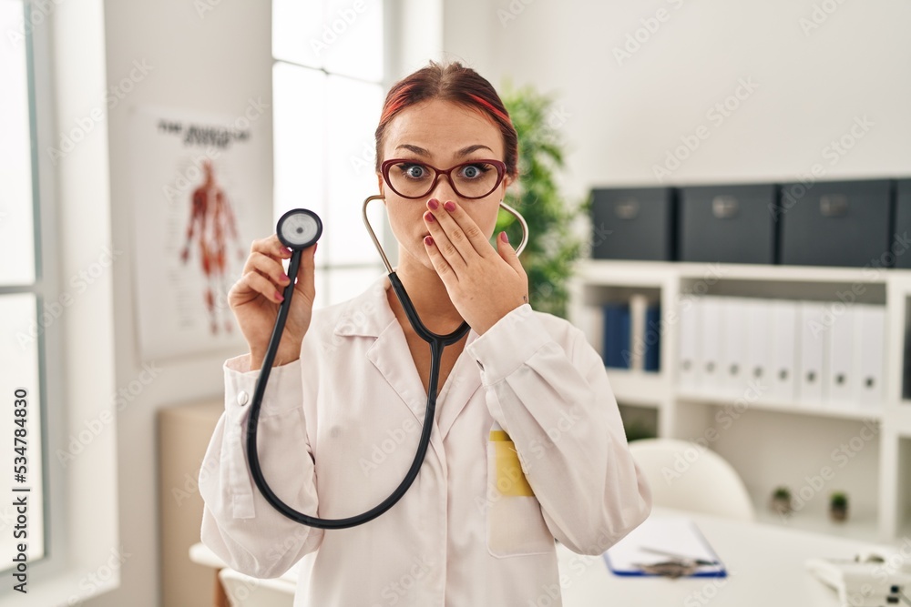 Young caucasian woman wearing doctor uniform using stethoscope covering mouth with hand, shocked and afraid for mistake. surprised expression Young caucasian woman wearing doctor uniform using stethoscope covering mouth with hand, shocked and afraid for mistake. surprised expression