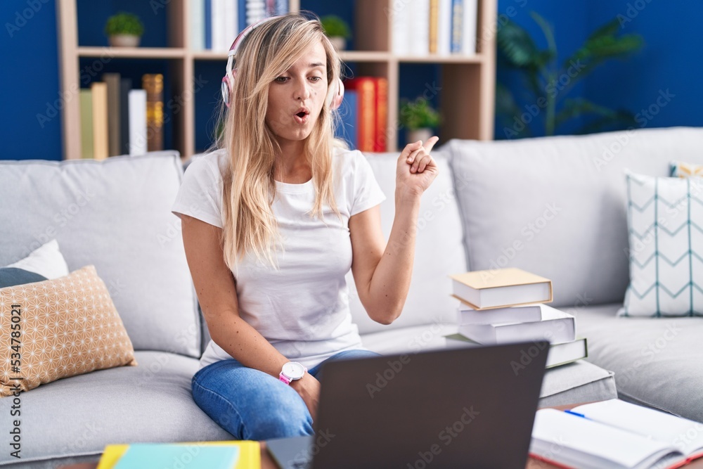 Young blonde woman studying using computer laptop at home surprised pointing with finger to the side, open mouth amazed expression. Young blonde woman studying using computer laptop at home surprised pointing with finger to the side, open mouth amazed expression.