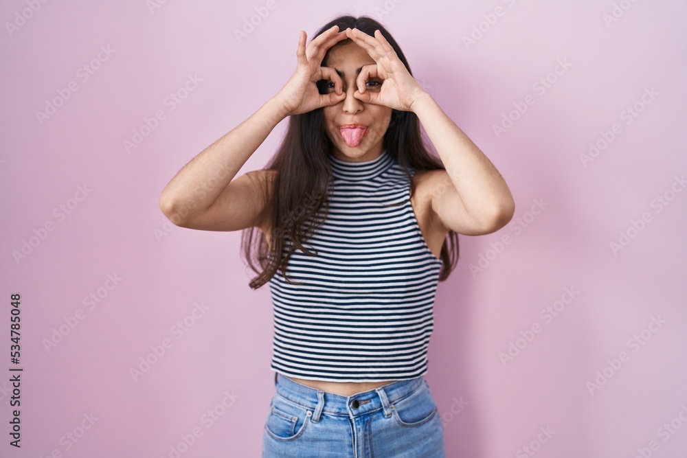 Young teenager girl wearing casual striped t shirt doing ok gesture like binoculars sticking tongue out, eyes looking through fingers. crazy expression. Young teenager girl wearing casual striped t shirt doing ok gesture like binoculars sticking tongue out, eyes looking through fingers. crazy expression.