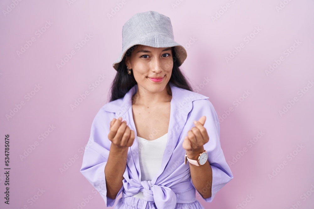 Young hispanic woman standing over pink background wearing hat doing money gesture with hands, asking for salary payment, millionaire business Young hispanic woman standing over pink background wearing hat doing money gesture with hands, asking for salary payment, millionaire business