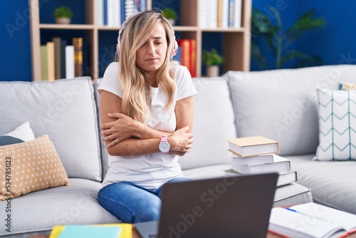 Young blonde woman studying using computer laptop at home shaking and freezing for winter cold with sad and shock expression on face