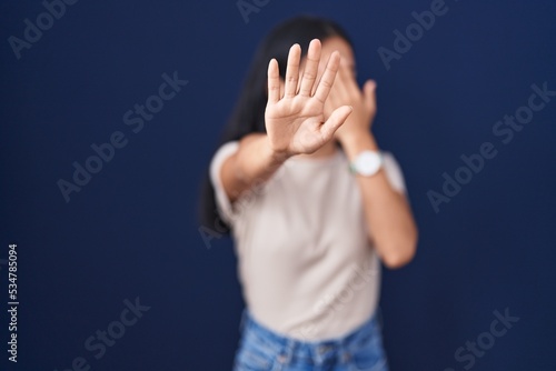 Young hispanic woman standing over blue background covering eyes with hands and doing stop gesture with sad and fear expression. embarrassed and negative concept.