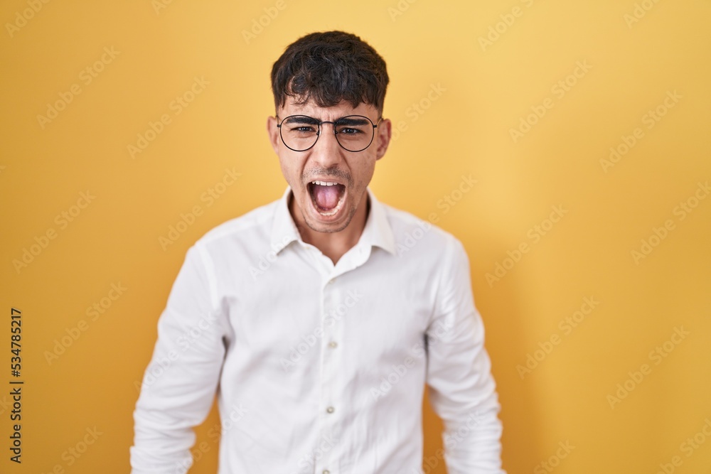 Young hispanic man standing over yellow background angry and mad screaming frustrated and furious, shouting with anger. rage and aggressive concept. Young hispanic man standing over yellow background angry and mad screaming frustrated and furious, shouting with anger. rage and aggressive concept.