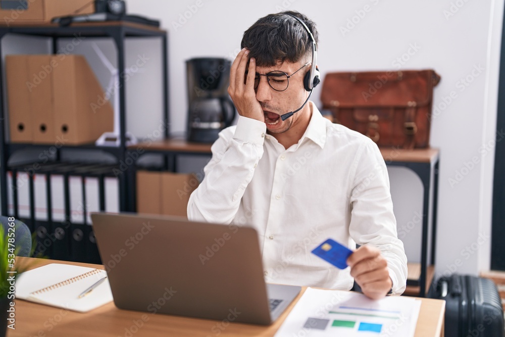 Young hispanic man working using computer laptop holding credit card yawning tired covering half face, eye and mouth with hand. face hurts in pain. Young hispanic man working using computer laptop holding credit card yawning tired covering half face, eye and mouth with hand. face hurts in pain.