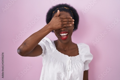 African woman with curly hair standing over pink background smiling and laughing with hand on face covering eyes for surprise. blind concept.