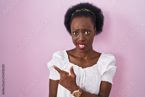 African woman with curly hair standing over pink background pointing aside worried and nervous with forefinger, concerned and surprised expression