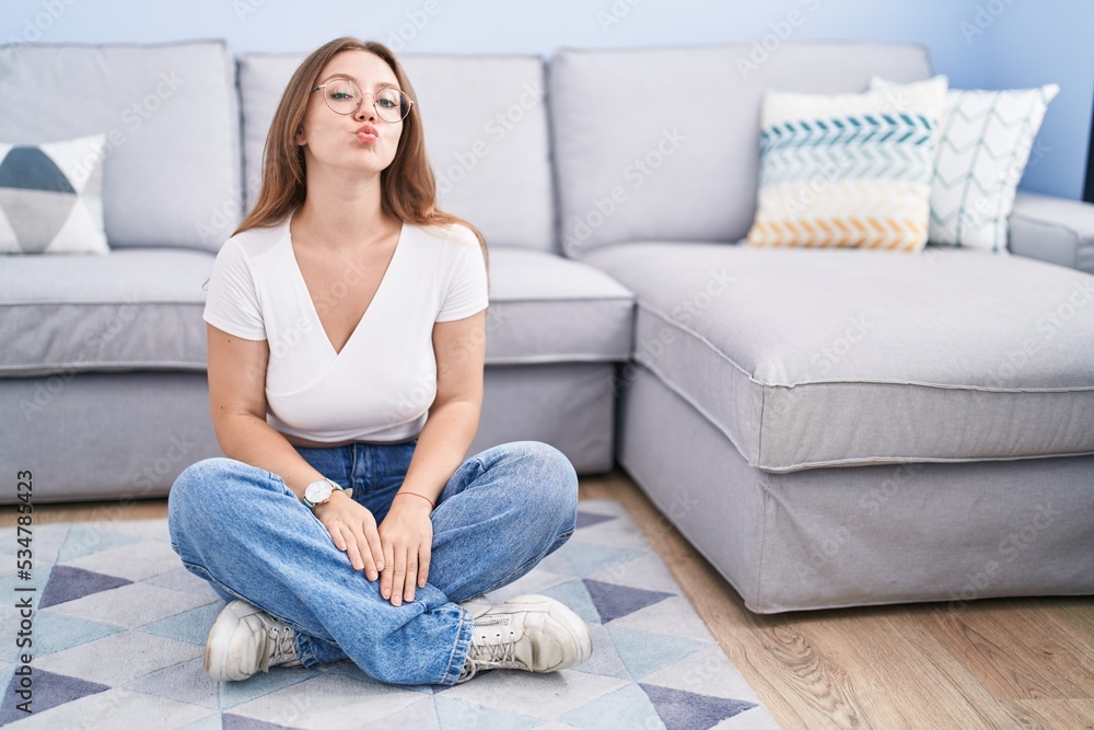 Young caucasian woman sitting on the floor at the living room looking at the camera blowing a kiss on air being lovely and sexy. love expression. Young caucasian woman sitting on the floor at the living room looking at the camera blowing a kiss on air being lovely and sexy. love expression.