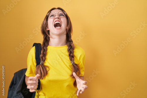 Young caucasian woman wearing student backpack over yellow background angry and mad screaming frustrated and furious, shouting with anger. rage and aggressive concept.