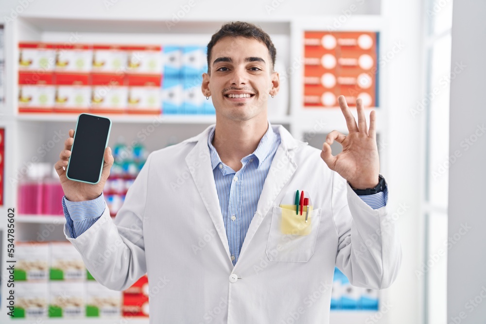 Handsome hispanic man working at pharmacy drugstore showing smartphone screen doing ok sign with fingers, smiling friendly gesturing excellent symbol