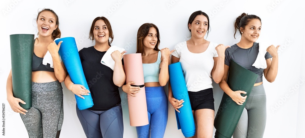 Group of women holding yoga mat standing over isolated background smiling with happy face looking and pointing to the side with thumb up. Group of women holding yoga mat standing over isolated background smiling with happy face looking and pointing to the side with thumb up.