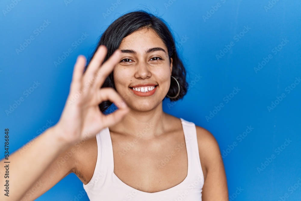 Young hispanic woman standing over blue background smiling positive doing ok sign with hand and fingers. successful expression. Young hispanic woman standing over blue background smiling positive doing ok sign with hand and fingers. successful expression.