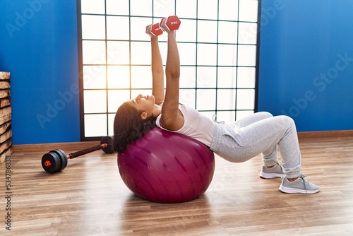 Young latin woman smiling confident training using dumbbells and fit ball at sport center