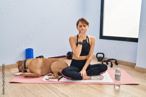 Young beautiful woman sitting on yoga mat bored yawning tired covering mouth with hand. restless and sleepiness.