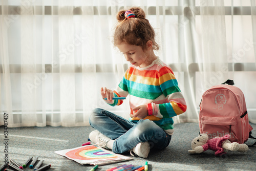 A cute little girl sits on the floor in the rays of the sun and draws a rainbow with colorful markers. She is dressed in a colored jumper. Creation. Childhood.