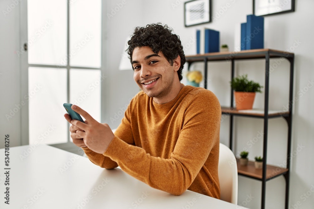 Young hispanic man using smartphone sitting on the table at home. Young hispanic man using smartphone sitting on the table at home.