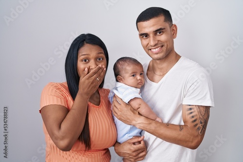 Young hispanic couple with baby standing together over isolated background laughing and embarrassed giggle covering mouth with hands, gossip and scandal concept