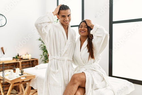 Young latin couple wearing towel standing at beauty center smiling confident touching hair with hand up gesture  posing attractive and fashionable