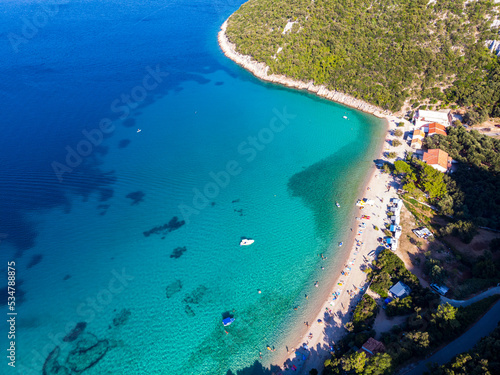 aerial view of a paradisiacal Mediterranean bay with turquoise water surrounded by massive mountains, with floating motorboats, with a small bushy island; the bay on the peljesac peninsula seen from a © Jakub