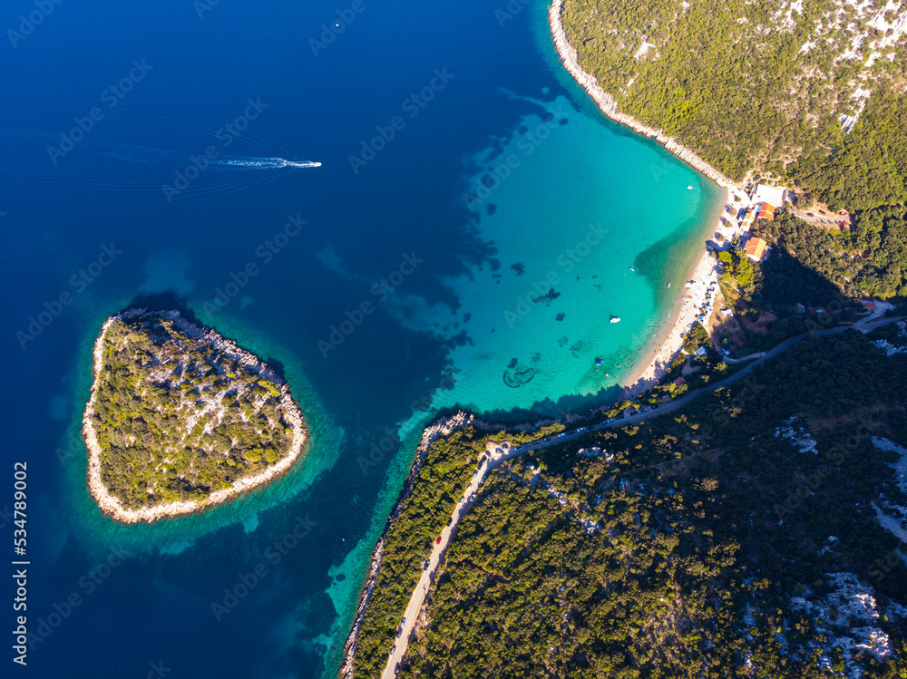 aerial view of a paradisiacal Mediterranean bay with turquoise water surrounded by massive mountains, with floating motorboats, with a small bushy island; the bay on the peljesac peninsula seen from a
