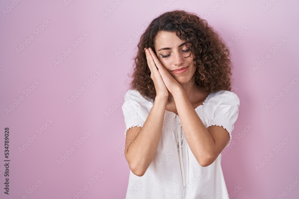 Hispanic woman with curly hair standing over pink background sleeping tired dreaming and posing with hands together while smiling with closed eyes.