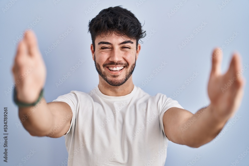 Hispanic man with beard standing over white background looking at the camera smiling with open arms for hug. cheerful expression embracing happiness. Hispanic man with beard standing over white background looking at the camera smiling with open arms for hug. cheerful expression embracing happiness.