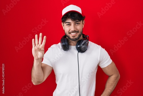 Hispanic man with beard wearing gamer hat and headphones showing and pointing up with fingers number four while smiling confident and happy.