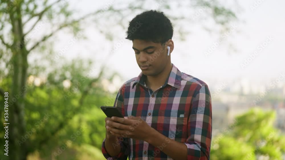 African American teenager in earphones texting on smartphone in park, leisure African American teenager in earphones texting on smartphone in park, leisure