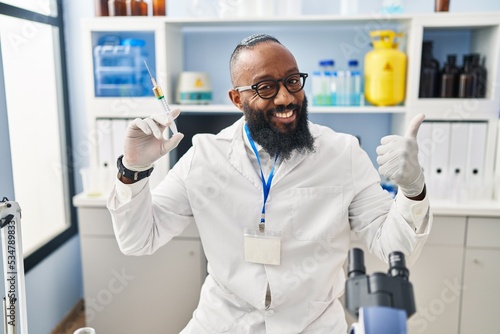 African american man working at scientist laboratory holding syringe smiling happy and positive  thumb up doing excellent and approval sign