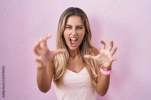 Young blonde woman standing over pink background shouting frustrated with rage, hands trying to strangle, yelling mad