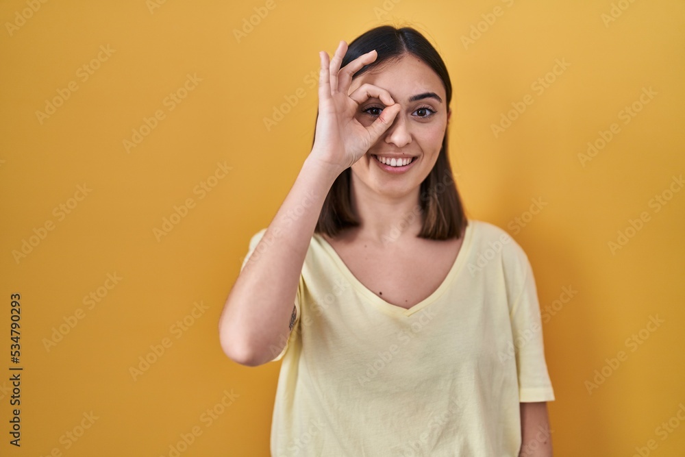 Hispanic girl wearing casual t shirt over yellow background doing ok gesture with hand smiling, eye looking through fingers with happy face. Hispanic girl wearing casual t shirt over yellow background doing ok gesture with hand smiling, eye looking through fingers with happy face.