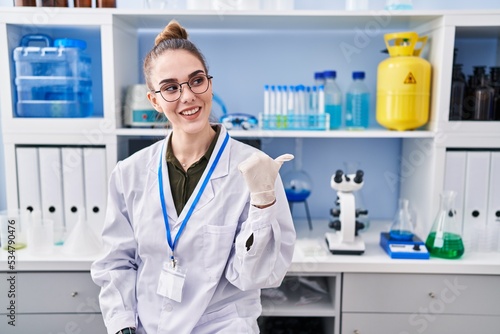 Young hispanic girl working at scientist laboratory pointing thumb up to the side smiling happy with open mouth