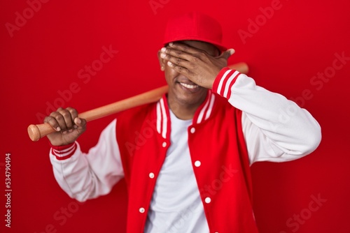 Young hispanic man playing baseball holding bat smiling and laughing with hand on face covering eyes for surprise. blind concept.