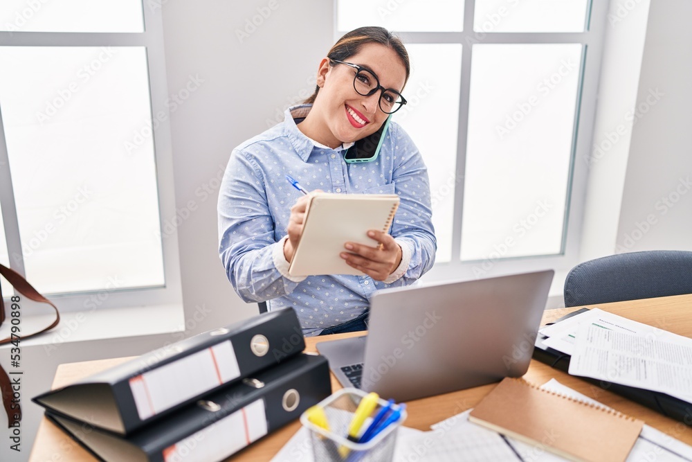 Young beautiful hispanic woman business worker talking on smartphone writing on notebook at office Young beautiful hispanic woman business worker talking on smartphone writing on notebook at office