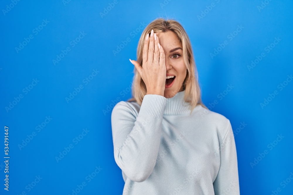 Young caucasian woman standing over blue background covering one eye with hand, confident smile on face and surprise emotion. Young caucasian woman standing over blue background covering one eye with hand, confident smile on face and surprise emotion.