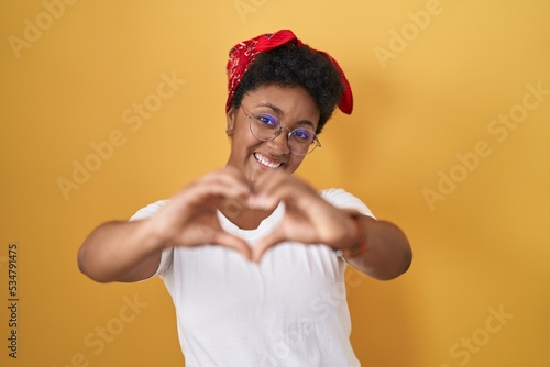 Young african american woman standing over yellow background smiling in love doing heart symbol shape with hands. romantic concept.