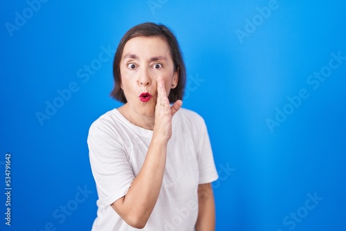 Middle age hispanic woman standing over blue background hand on mouth telling secret rumor, whispering malicious talk conversation