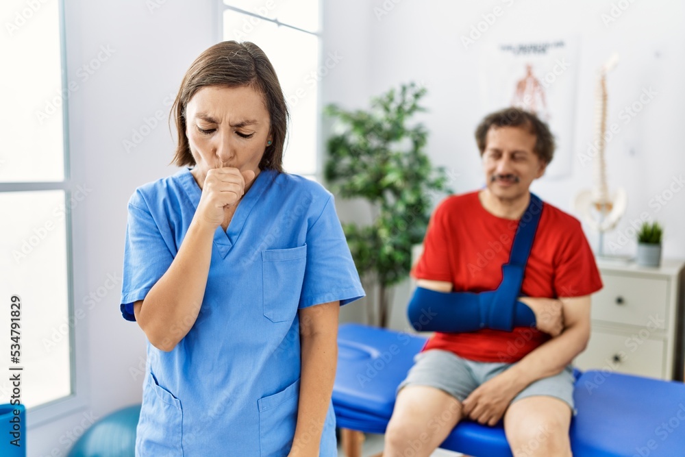 Obraz premium Middle age doctor woman with patient with arm injury at rehabilitation clinic feeling unwell and coughing as symptom for cold or bronchitis. health care concept.