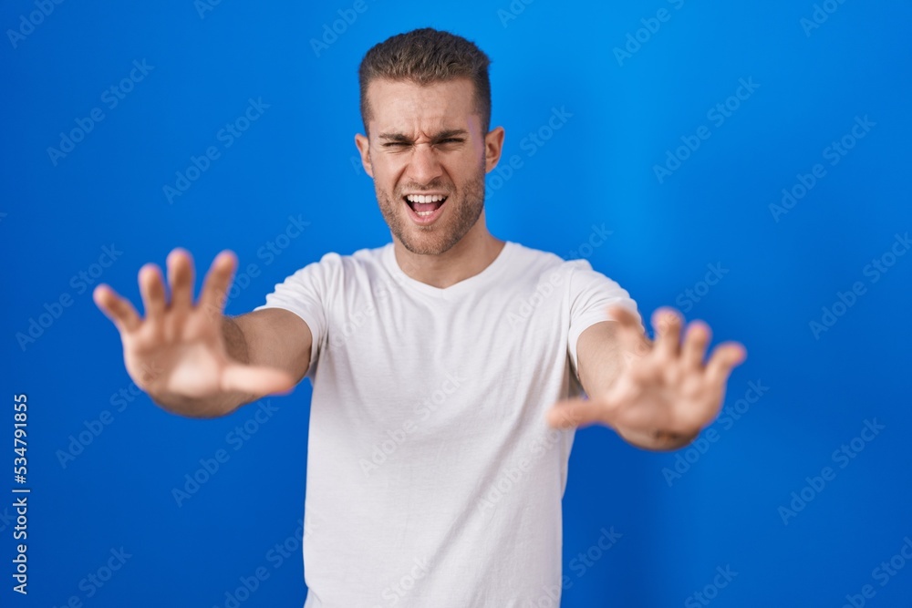 Young caucasian man standing over blue background afraid and terrified with fear expression stop gesture with hands, shouting in shock. panic concept. Young caucasian man standing over blue background afraid and terrified with fear expression stop gesture with hands, shouting in shock. panic concept.