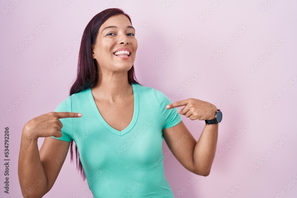 Young hispanic woman standing over pink background looking confident with smile on face, pointing oneself with fingers proud and happy.