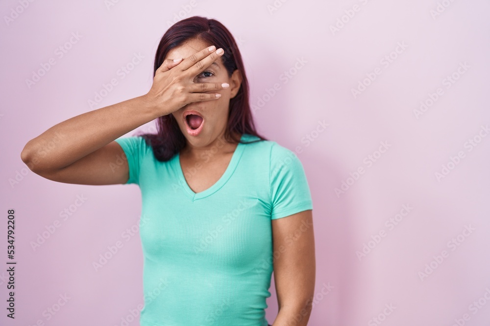 Young hispanic woman standing over pink background peeking in shock covering face and eyes with hand, looking through fingers with embarrassed expression. Young hispanic woman standing over pink background peeking in shock covering face and eyes with hand, looking through fingers with embarrassed expression.