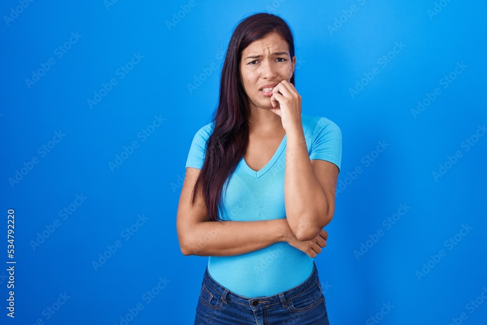 Young hispanic woman standing over blue background looking stressed and nervous with hands on mouth biting nails. anxiety problem. Young hispanic woman standing over blue background looking stressed and nervous with hands on mouth biting nails. anxiety problem.