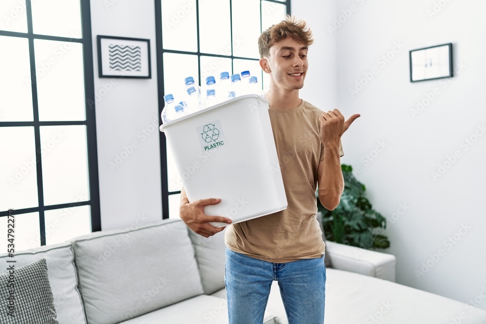 Young caucasian man holding wastebasket with recycling plastic bottles at home pointing thumb up to the side smiling happy with open mouth Young caucasian man holding wastebasket with recycling plastic bottles at home pointing thumb up to the side smiling happy with open mouth