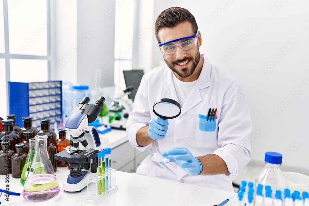 Young hispanic man wearing scientist uniform using loupe at laboratory