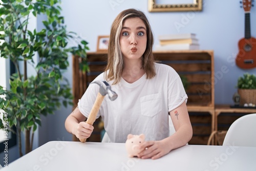 Blonde caucasian woman holding piggy bank and hammer puffing cheeks with funny face. mouth inflated with air, catching air.