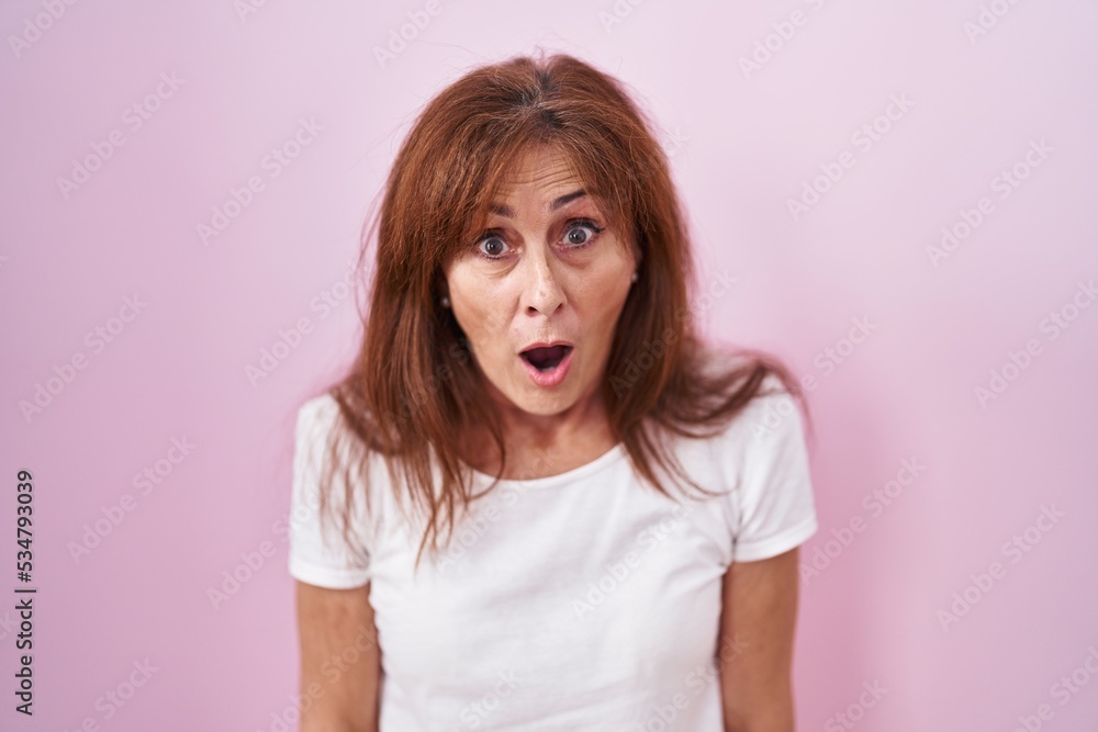 Middle age woman standing over pink background afraid and shocked with surprise and amazed expression, fear and excited face. Middle age woman standing over pink background afraid and shocked with surprise and amazed expression, fear and excited face.