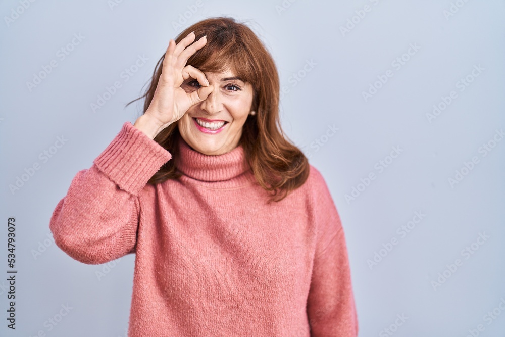 Middle age hispanic woman standing over isolated background doing ok gesture with hand smiling, eye looking through fingers with happy face. Middle age hispanic woman standing over isolated background doing ok gesture with hand smiling, eye looking through fingers with happy face.