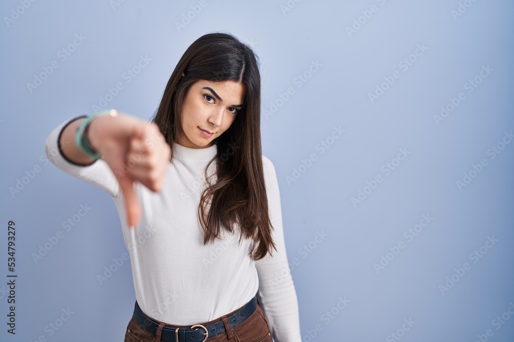 Young brunette woman standing over blue background looking unhappy and angry showing rejection and negative with thumbs down gesture. bad expression. Young brunette woman standing over blue background looking unhappy and angry showing rejection and negative with thumbs down gesture. bad expression.