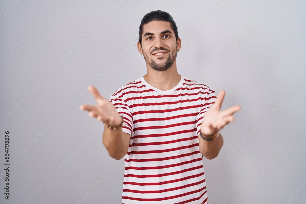 Hispanic man with long hair standing over isolated background smiling cheerful offering hands giving assistance and acceptance. Hispanic man with long hair standing over isolated background smiling cheerful offering hands giving assistance and acceptance.